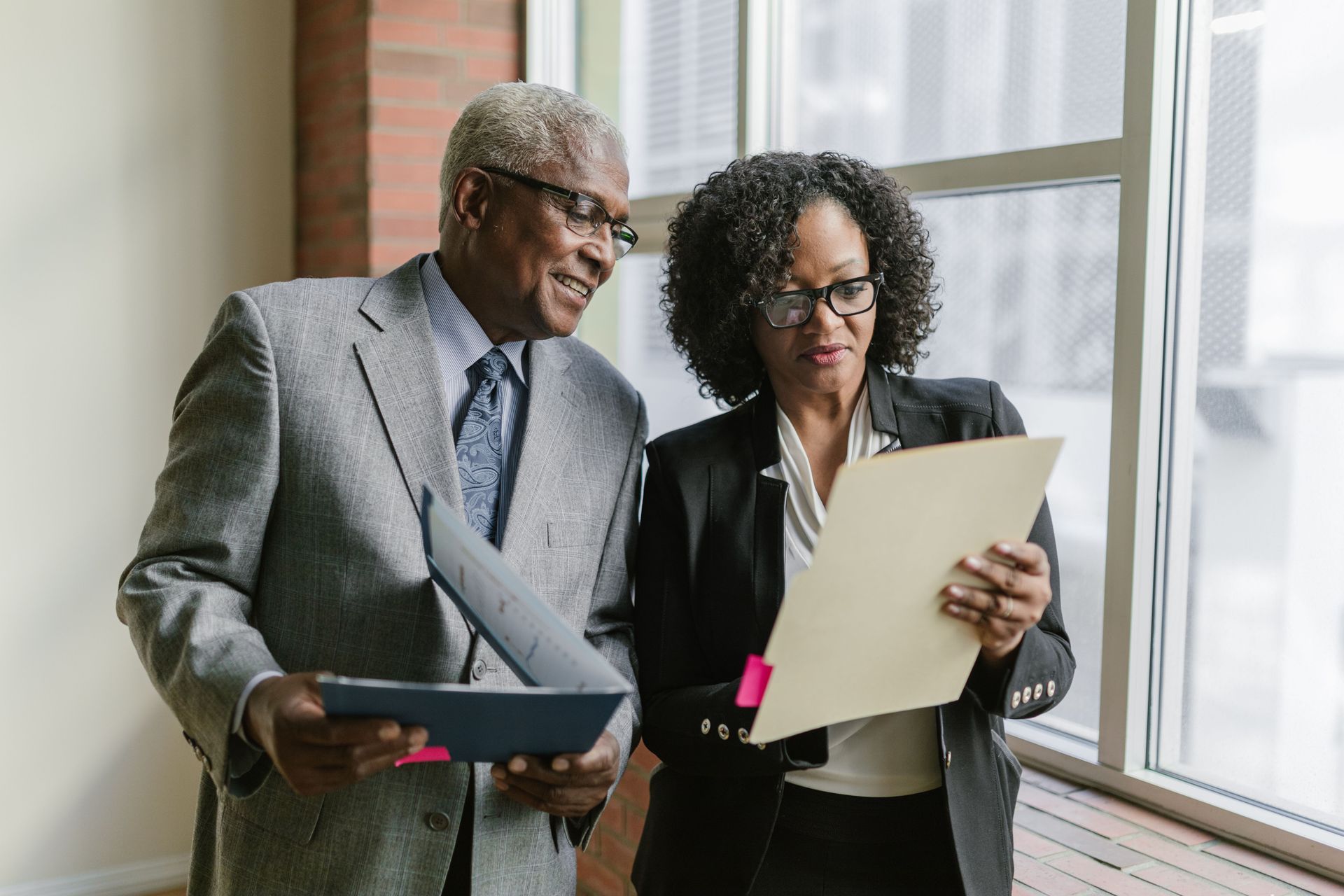 Two office workers reviewing documents by a window in a bright office