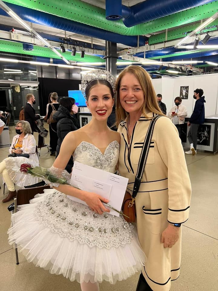 A ballet dancer in white tutu and a woman in beige coat smile for a photo. They stand indoors.
