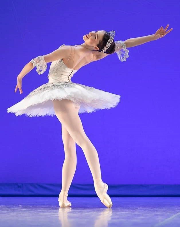 Ballerina in white tutu, arched back, arms extended, on pointes, on stage against purple backdrop.