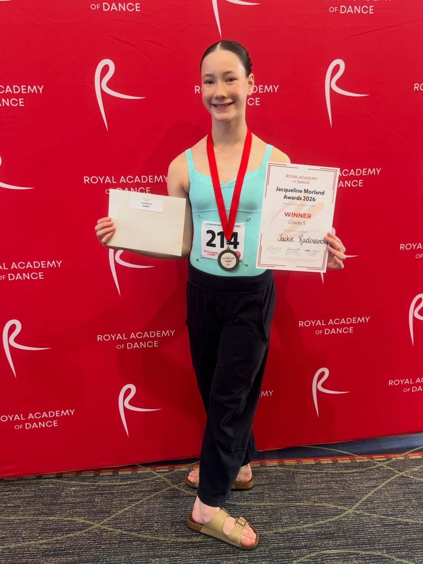 Young dancer holding a certificate and medal in front of a red Royal Academy of Dance backdrop