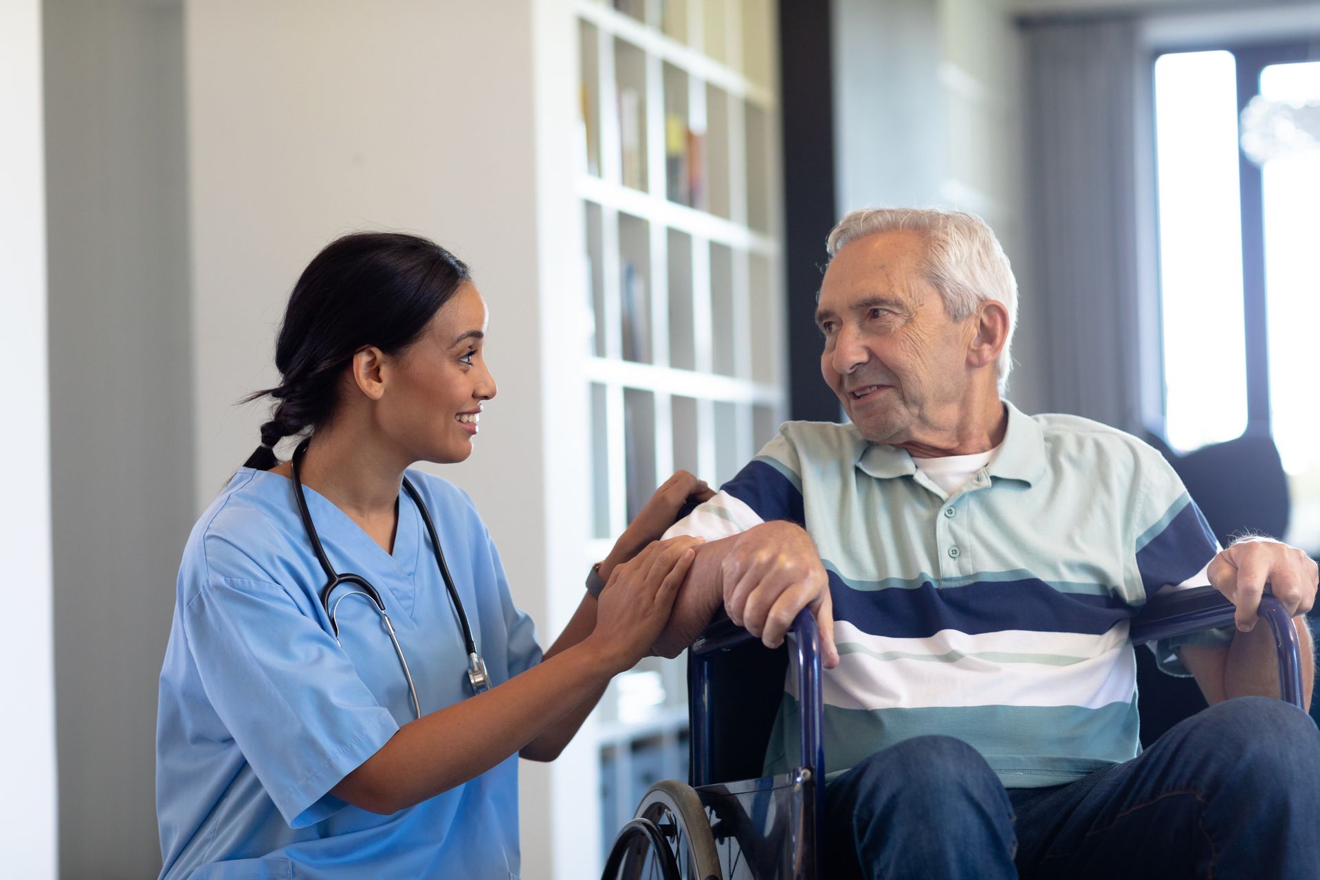Caregiver in blue scrubs with stethoscope, comforts a person in a wheelchair, smiling. Interior setting.