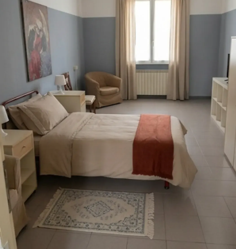 Camera da letto luminosa con letto singolo, sedia beige, finestra e pareti grigio chiaro in stile minimalista.