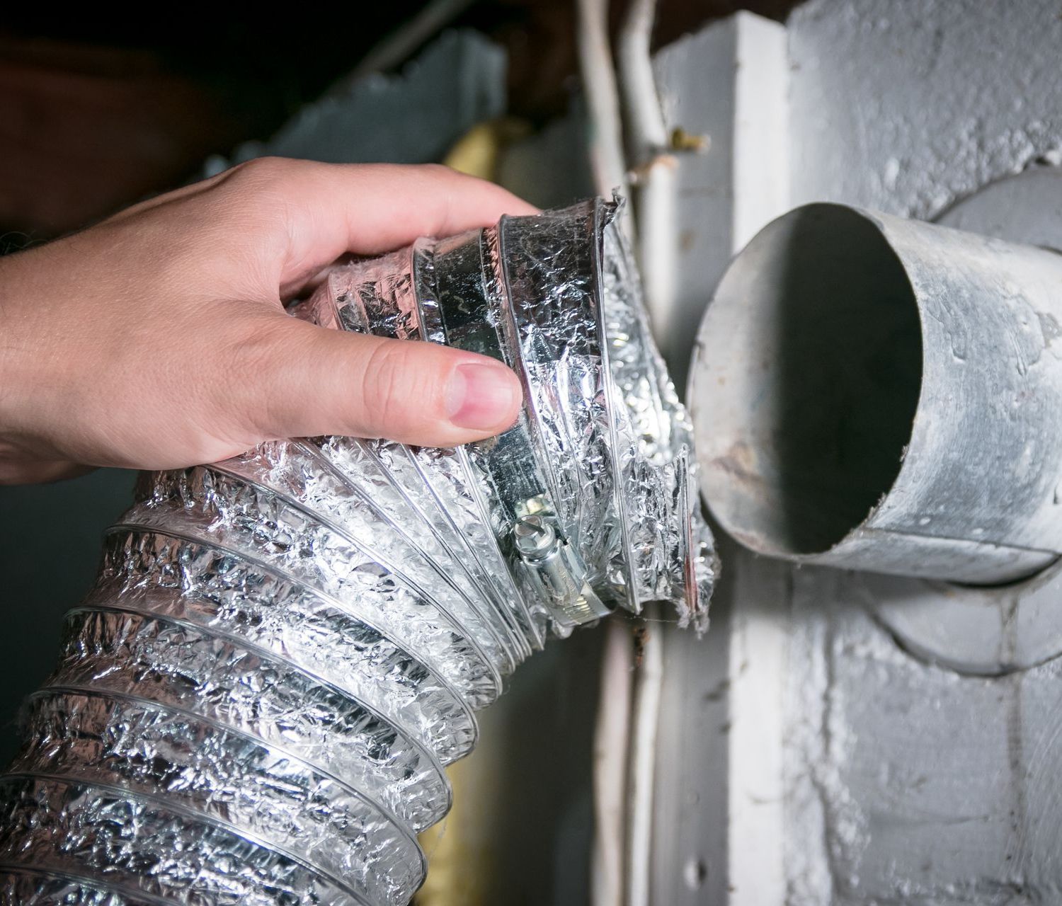 Hand holding a flexible silver duct hose near a wall vent opening