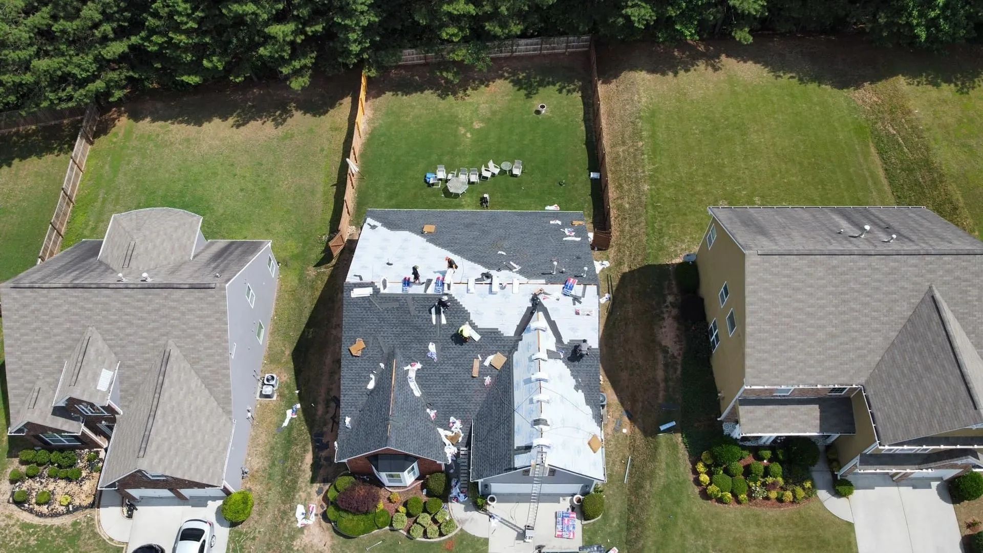 Aerial view of a residential house under construction with roof shingles being replaced, surrounded by neighboring homes.