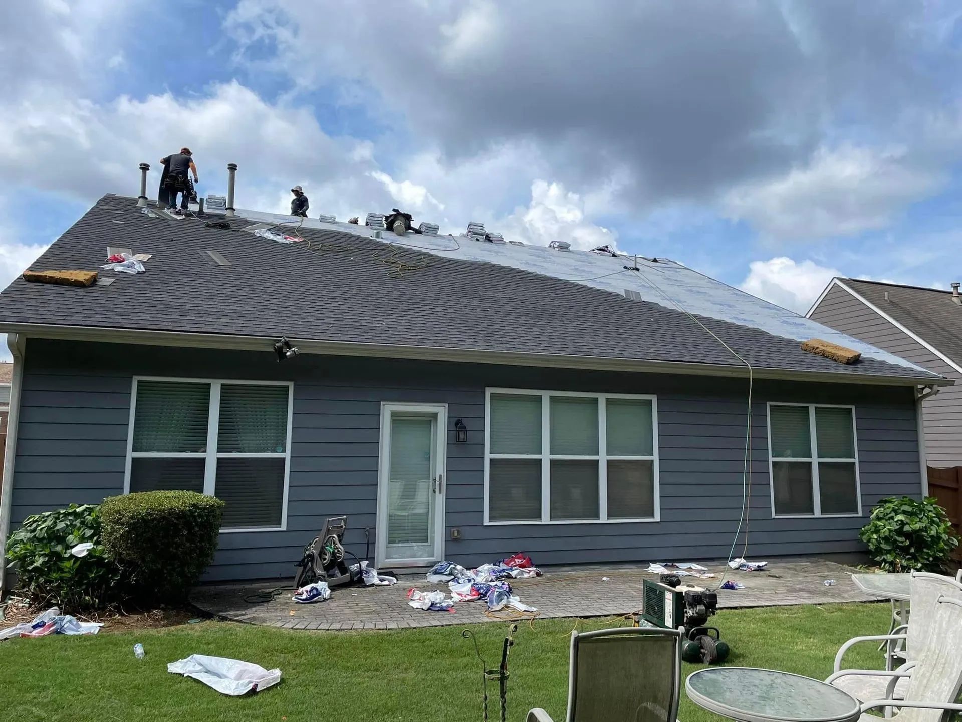 Workers on a roof installing black underlayment on a gray house after storm damage, with debris in the yard.