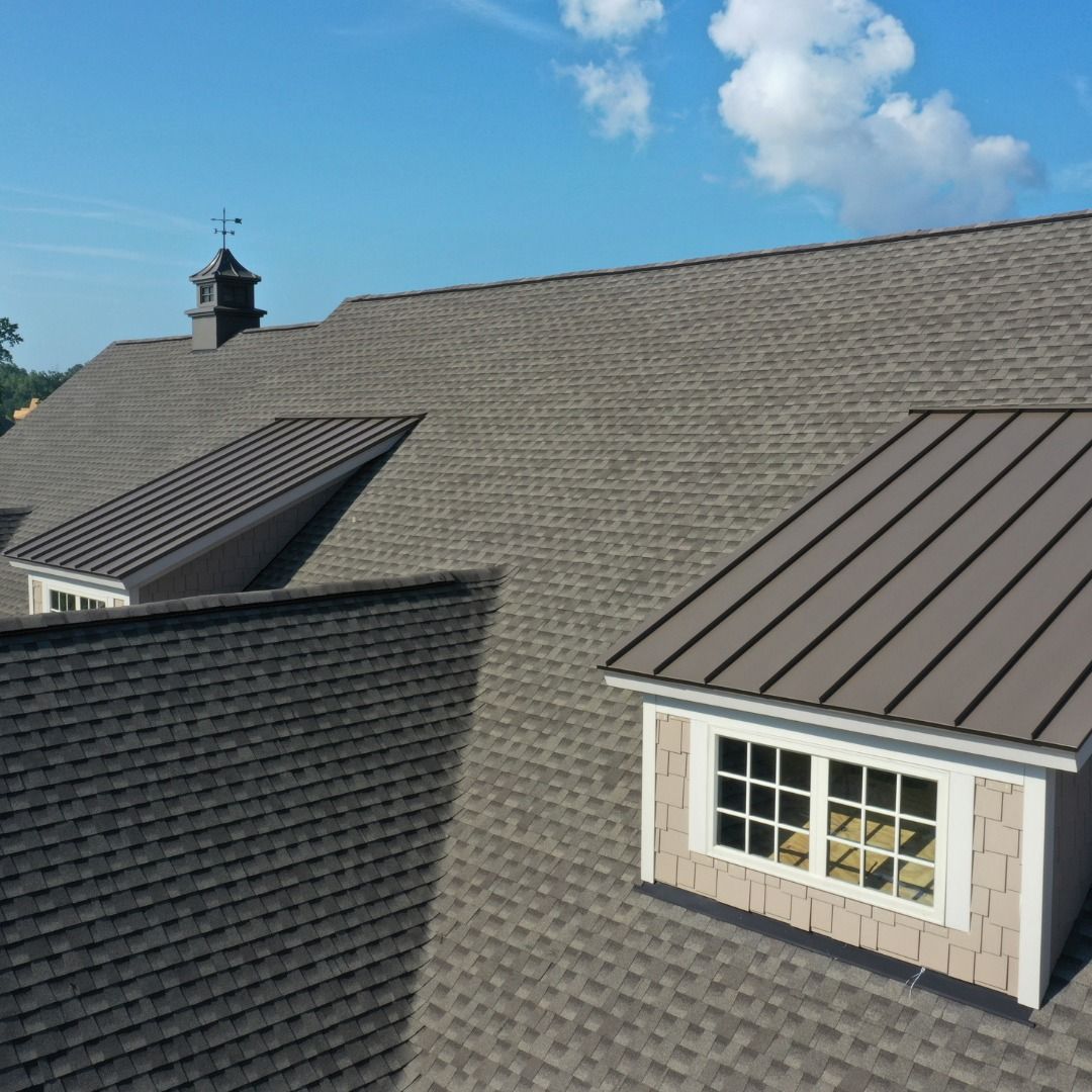 A roof with brown shingles and a cupola against a blue sky. Two metal-roofed dormers are visible.