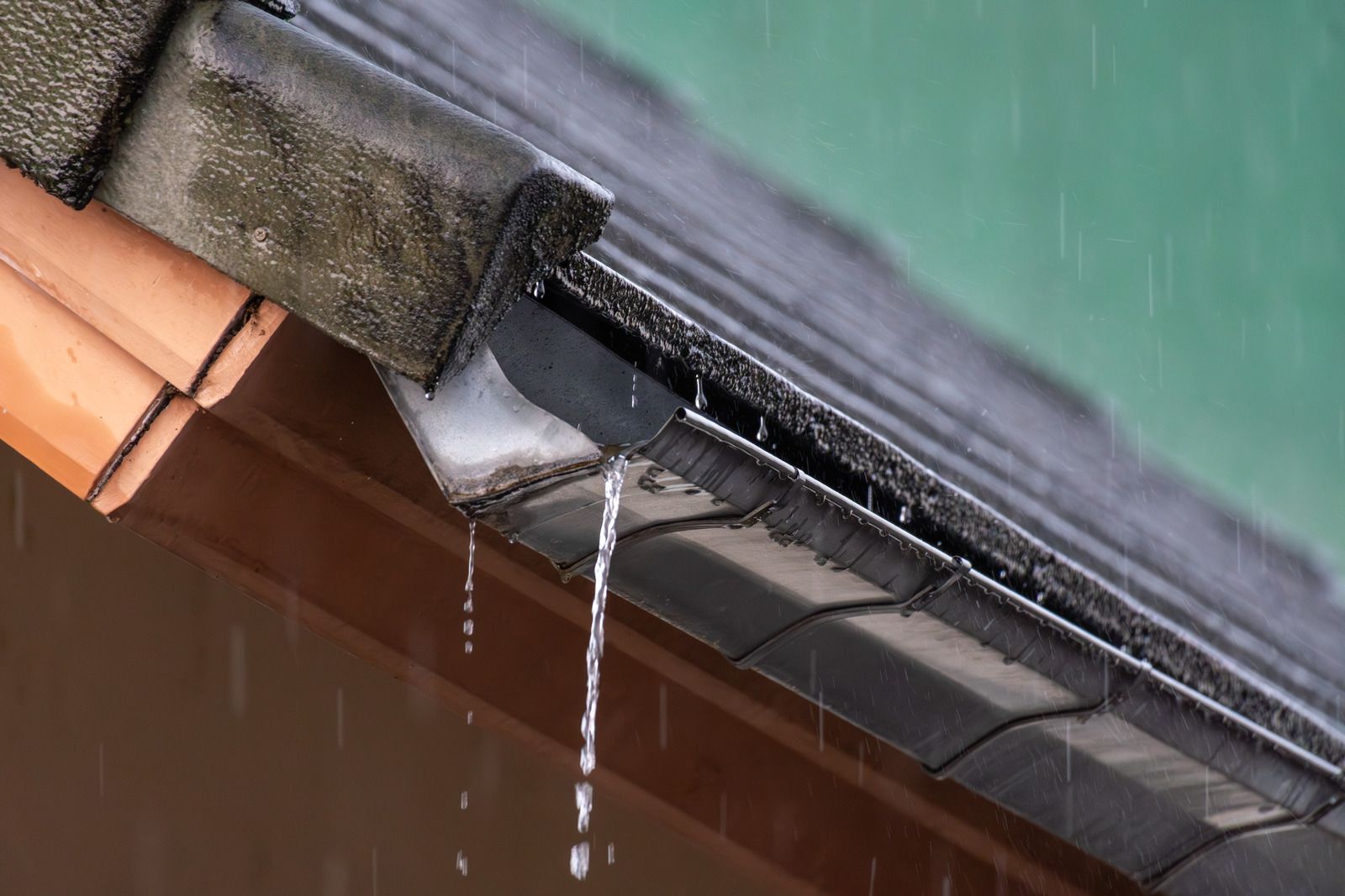 Rainwater dripping from a roof gutter beside terracotta tiles and a green wall