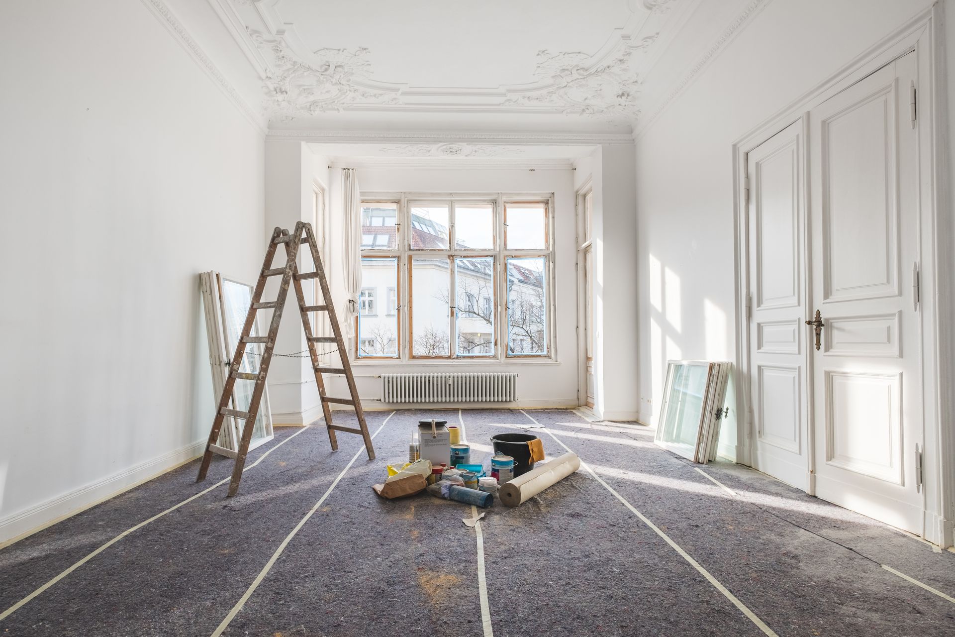 Empty room undergoing renovation; ladder, tools, and materials on the floor near window and double doors.