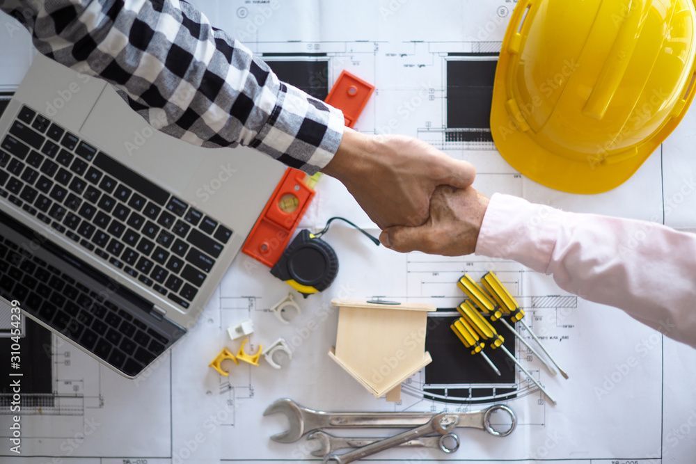 Two people shaking hands over blueprints with tools, laptop, and yellow hard hat.