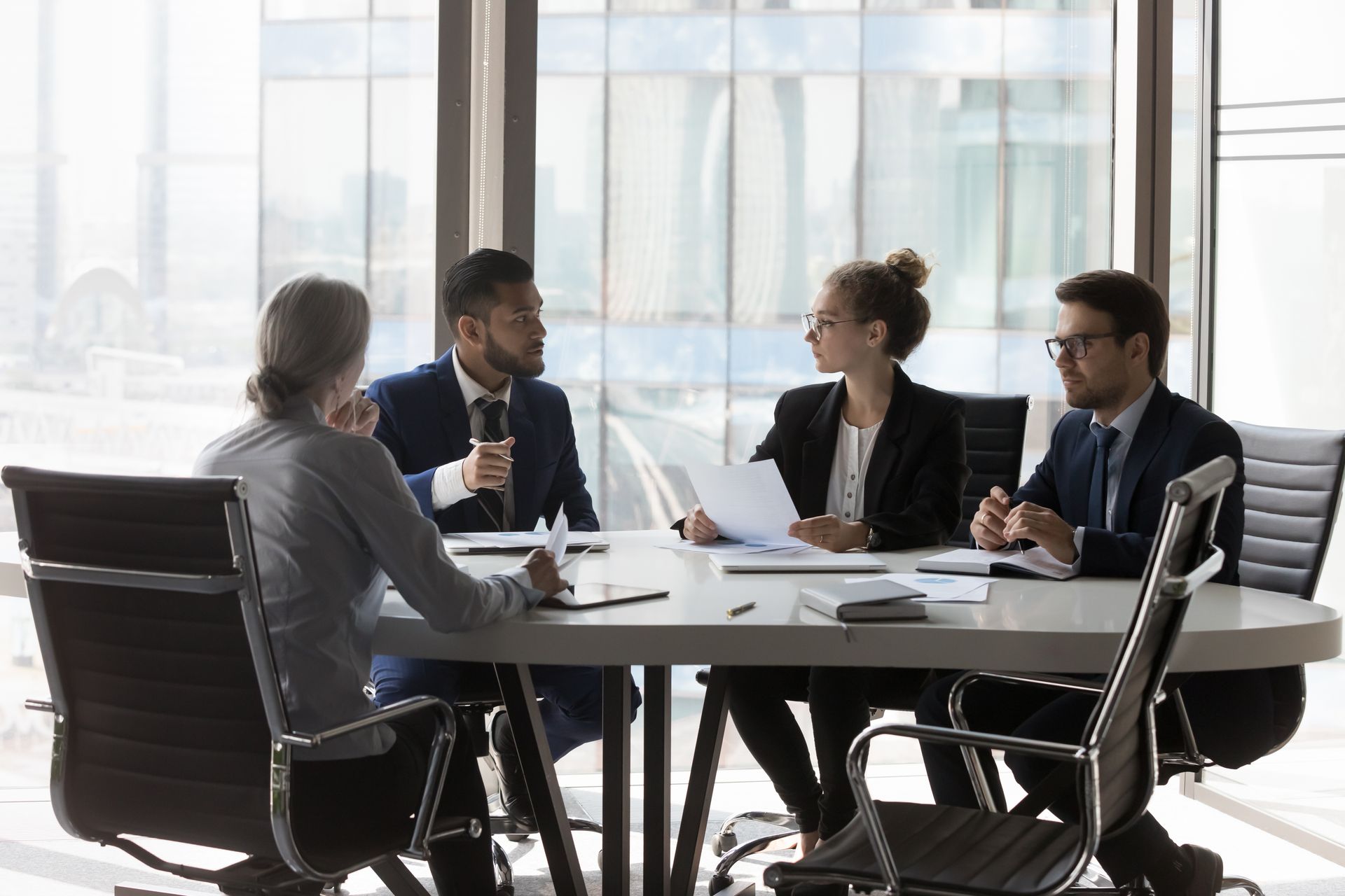 Four businesspeople in a modern office at a table, discussing documents.