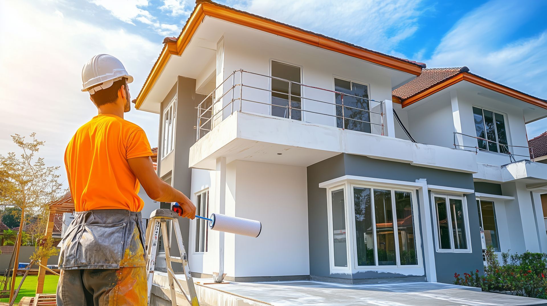 Painter in orange shirt and white hard hat painting a two-story house exterior gray and white.