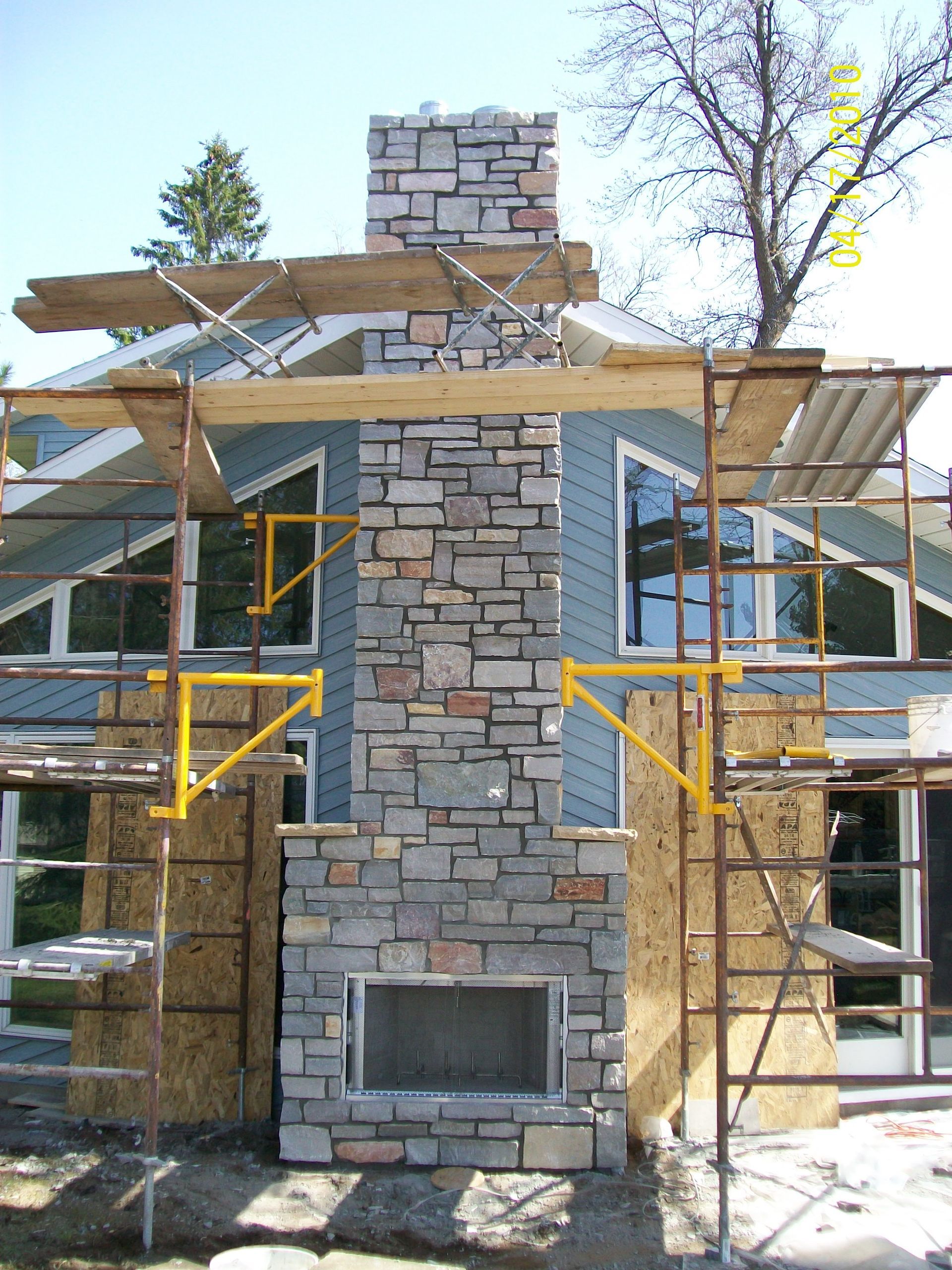 Stone chimney under construction with scaffolding on both sides and a house behind it
