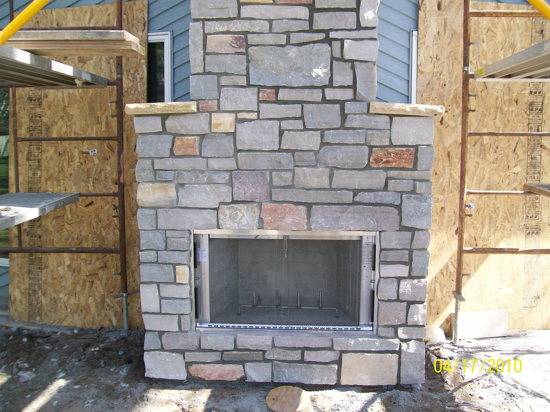 Stone fireplace under construction with a metal firebox opening, framed by plywood walls and scaffolding.
