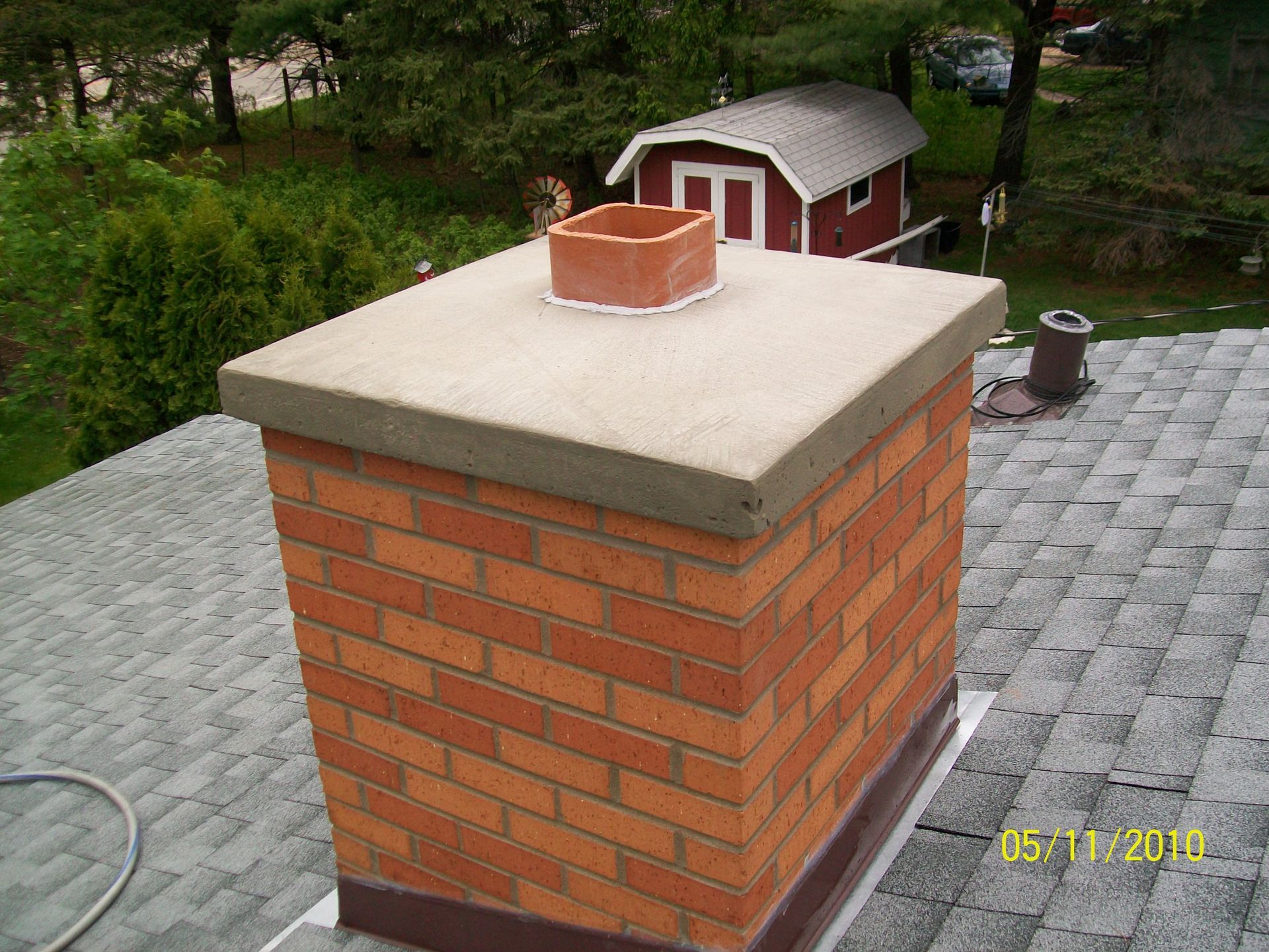 Brick chimney with a concrete cap on a gray shingled roof, with a small red shed in the background.