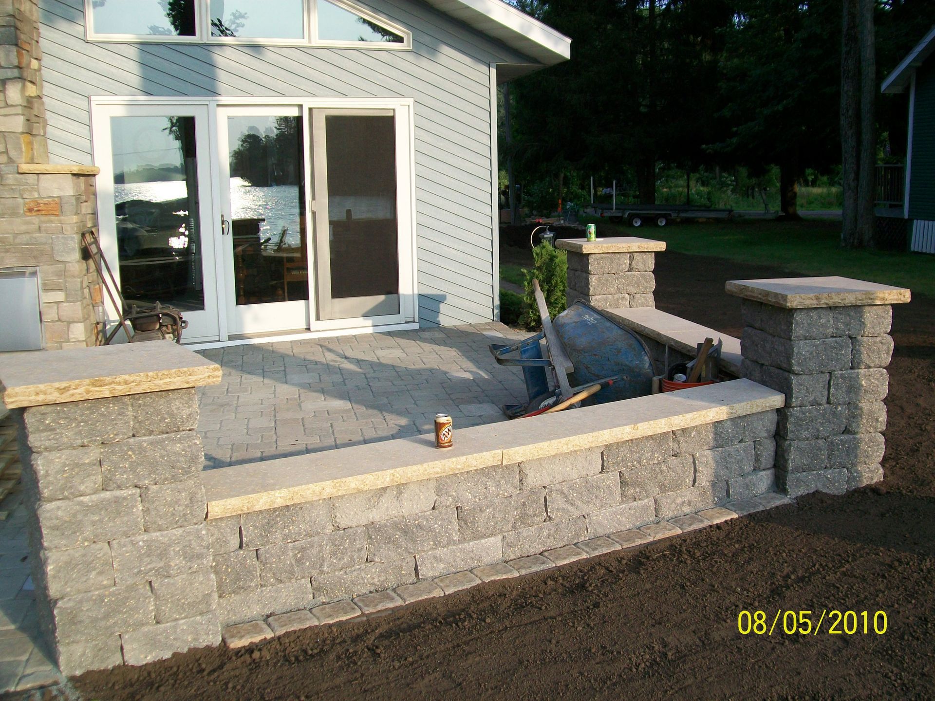 Stone patio and low retaining wall beside a house with sliding glass doors and steps to a yard.