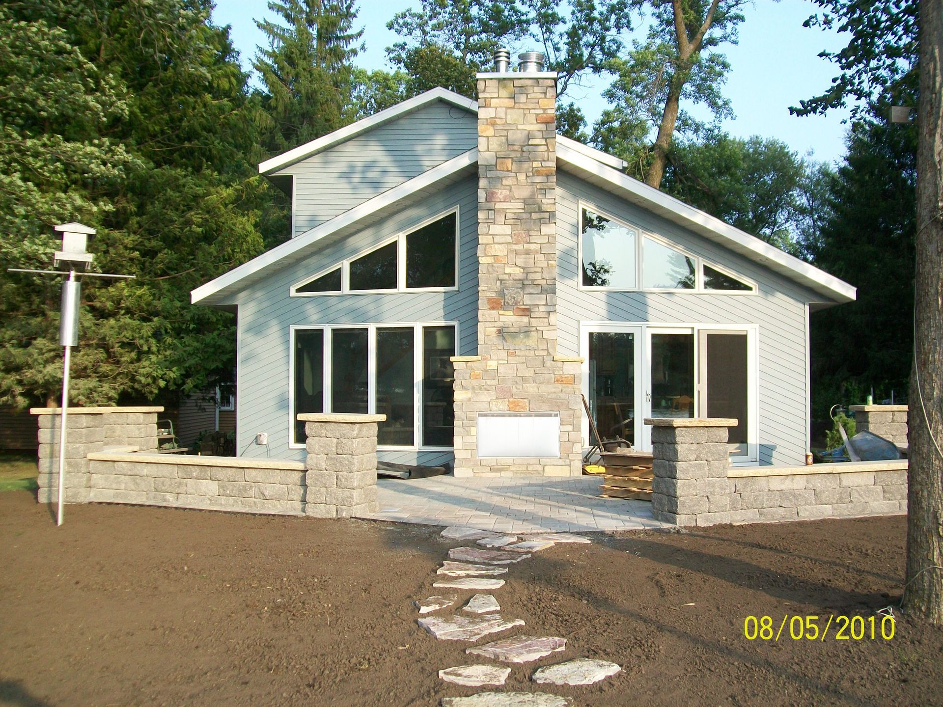 Modern house with stone chimney, large windows, and a front stone patio in a wooded setting