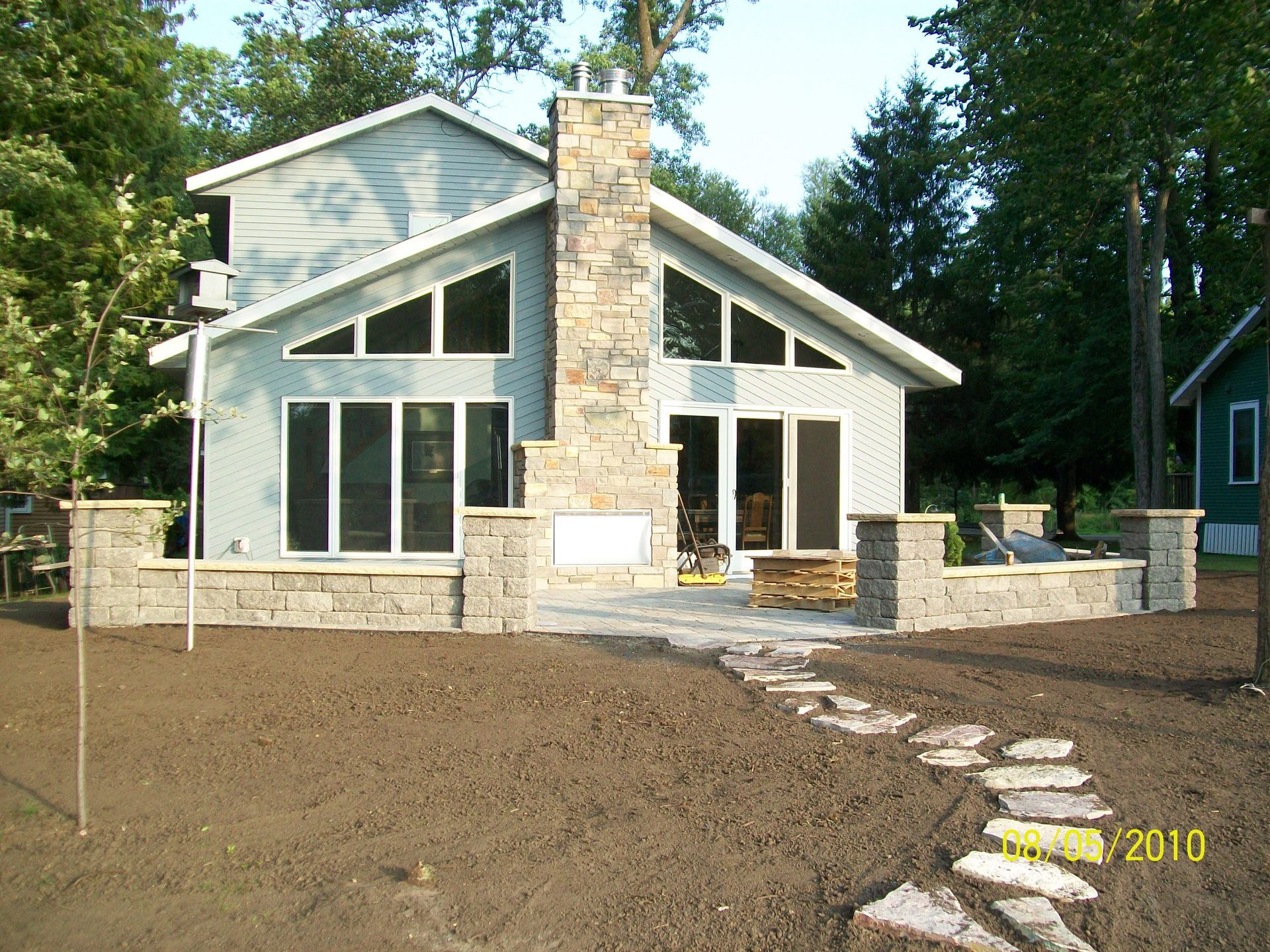 Modern cabin-style house with stone chimney, large windows, and a gravel yard with stone path