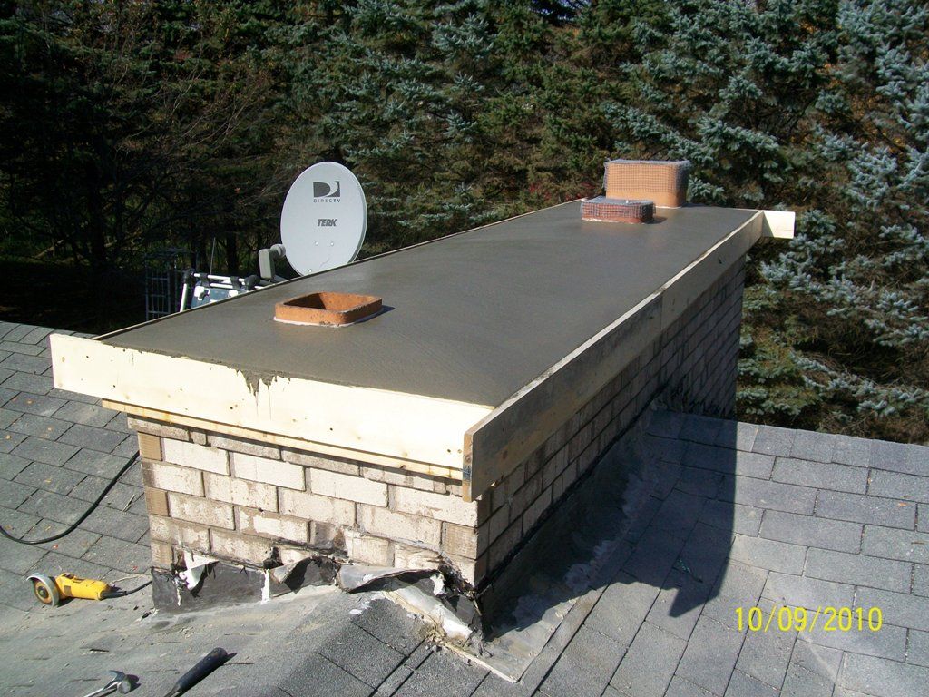 Rooftop with a brick chimney, metal cap, and satellite dish against trees and skylights.
