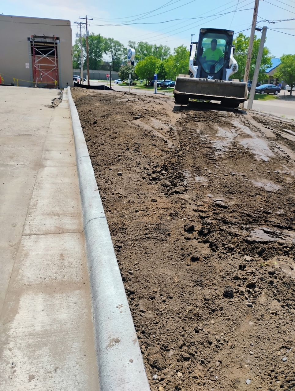 Construction excavator grading muddy ground beside a sidewalk and curb near a building.