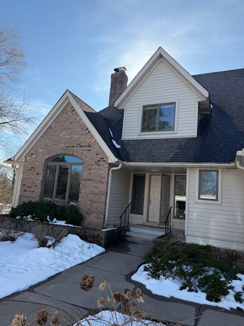 Snow-covered suburban house with brick and white siding, front porch, and blue roof under a clear sky