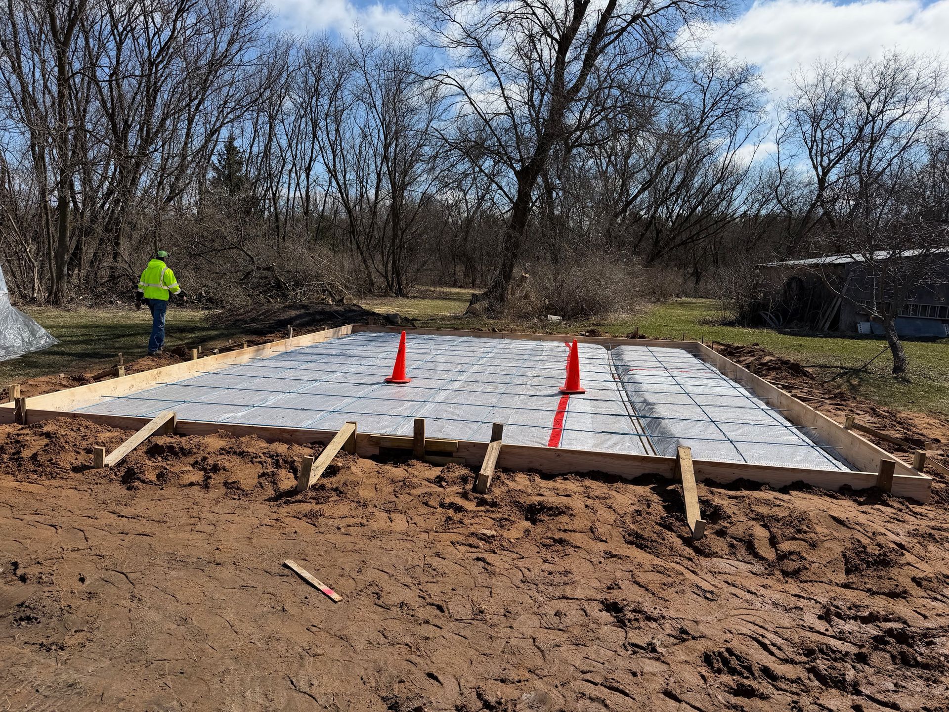 Construction site with concrete foundation slab, orange cones, and worker in a neon jacket
