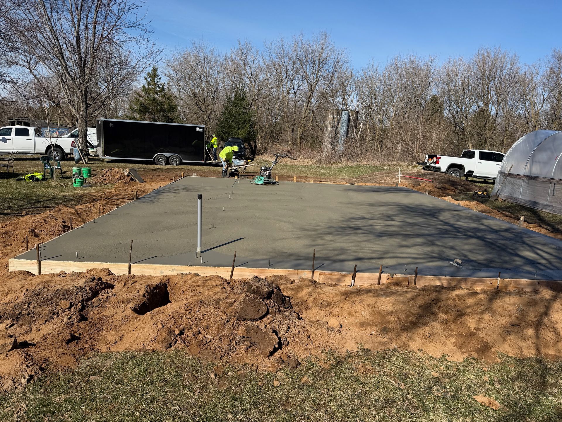 Concrete foundation slab being poured at a construction site, with workers, trucks, and bare trees nearby.