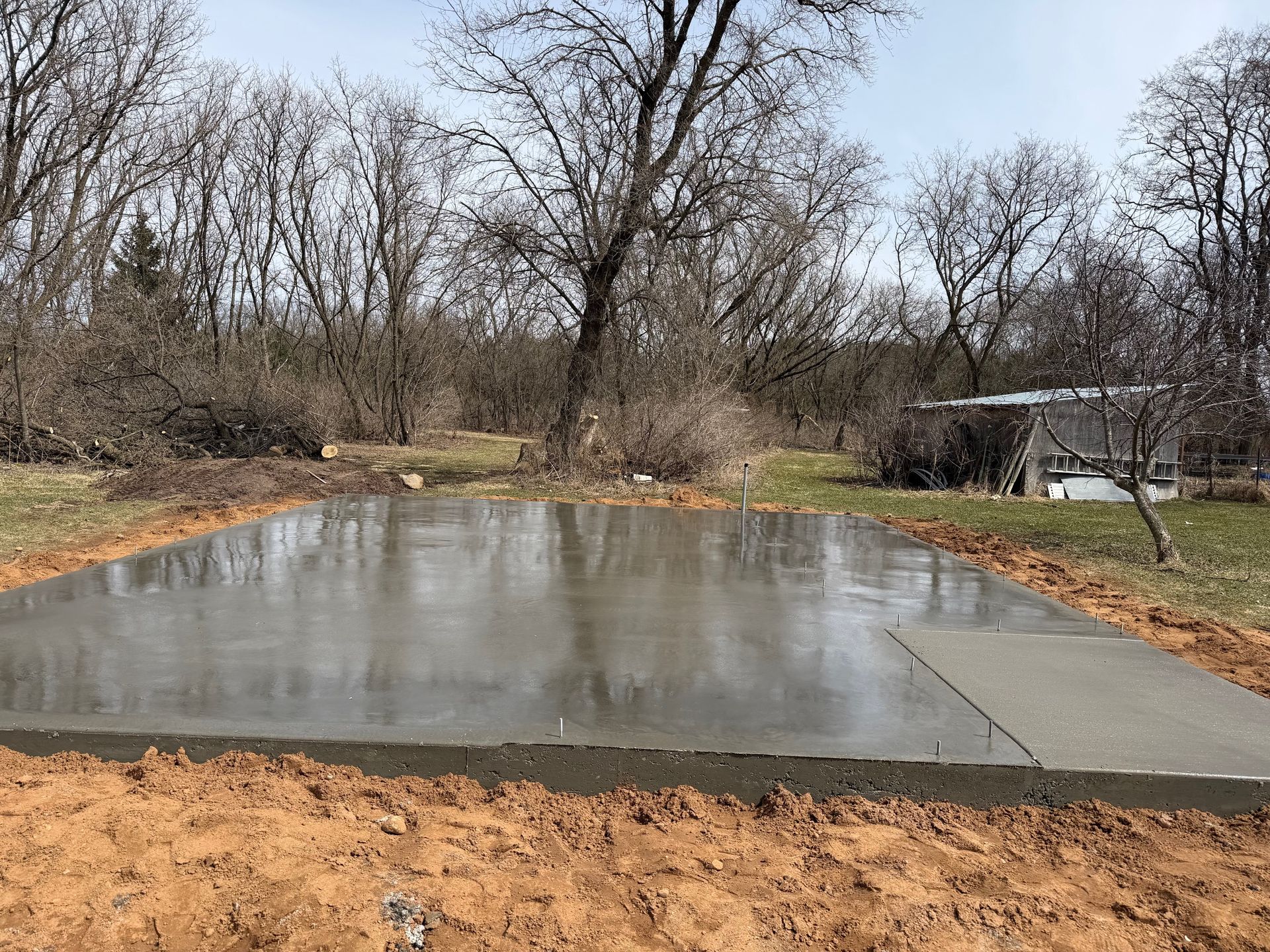 Fresh concrete slab in a muddy yard with leafless trees and a small shed in the background