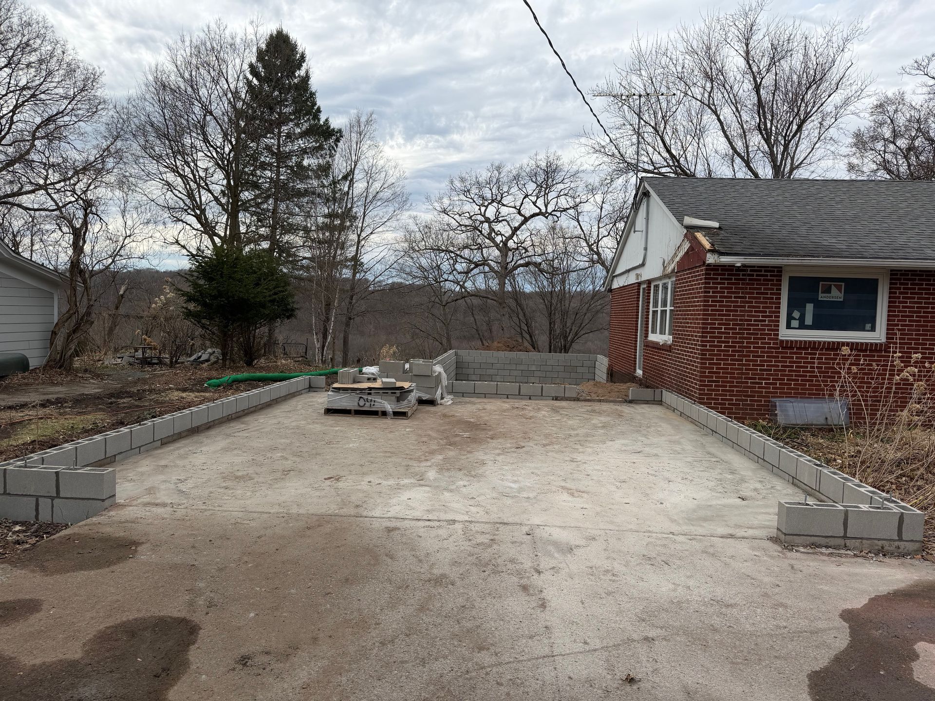 Backyard with a concrete patio, cinder block border, brick house, and trees under a cloudy sky