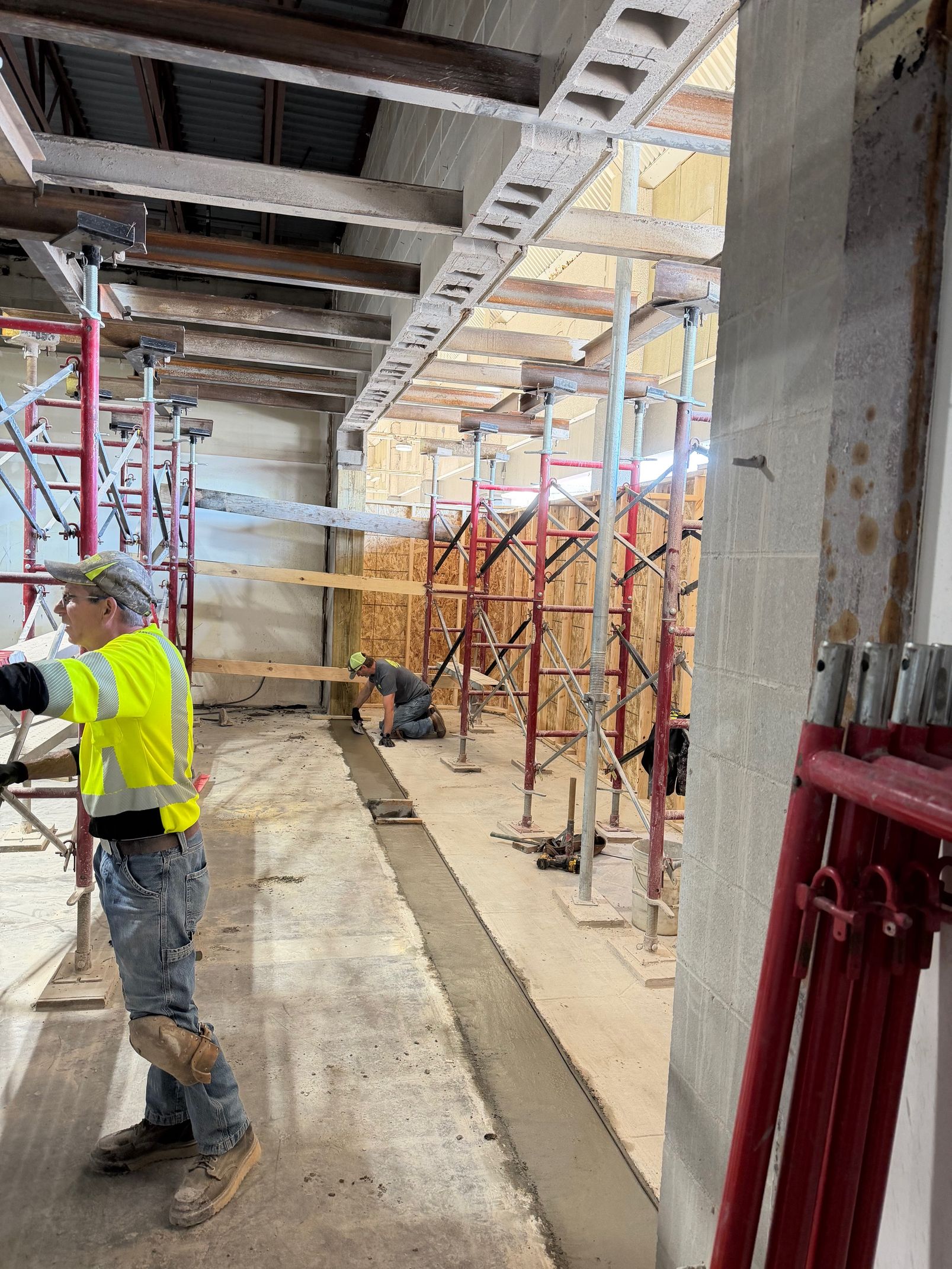 Construction worker in a yellow safety vest at a scaffolded indoor worksite.