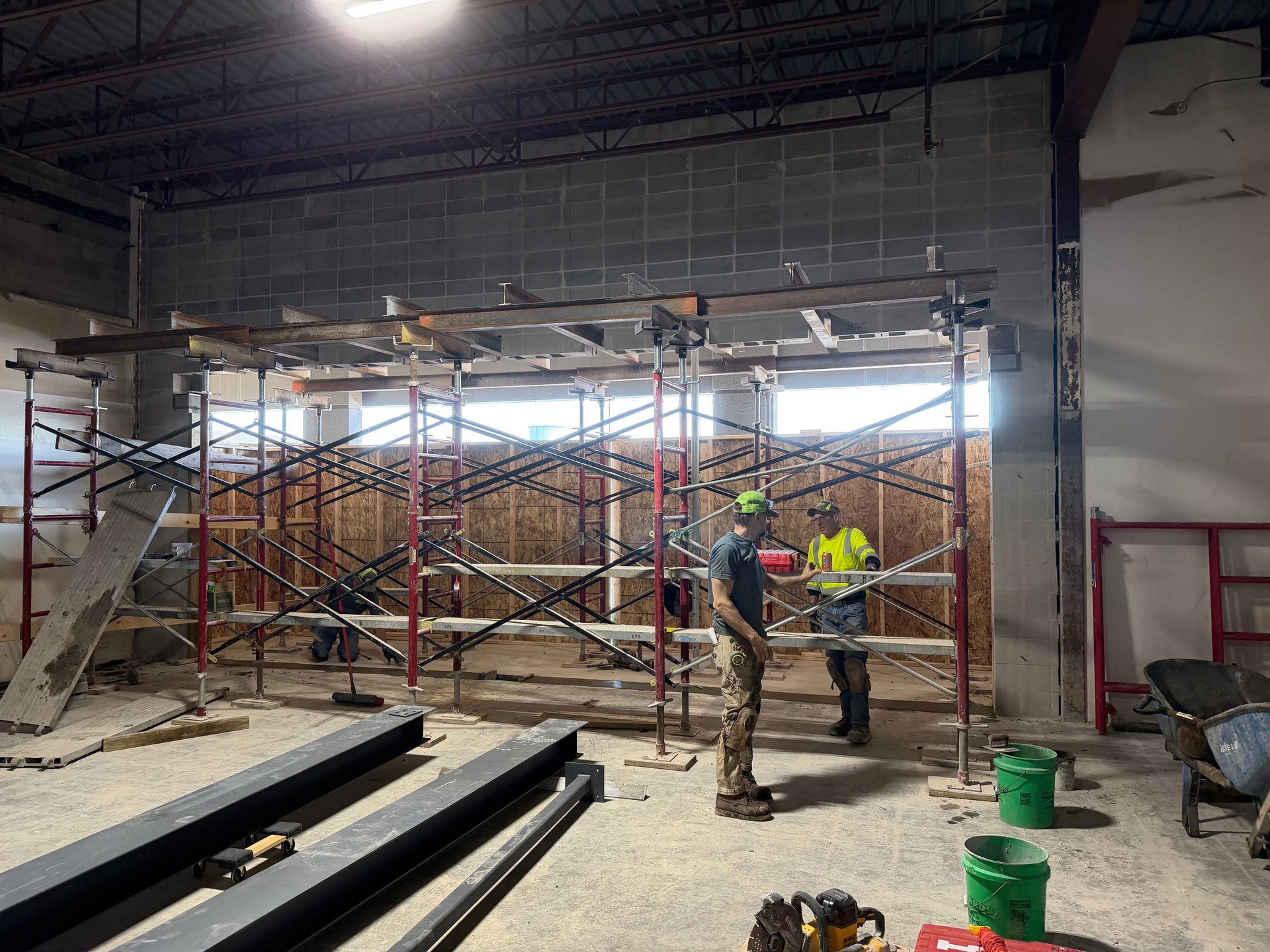 Workers on scaffolding inside an unfinished warehouse, with metal beams and construction materials nearby.