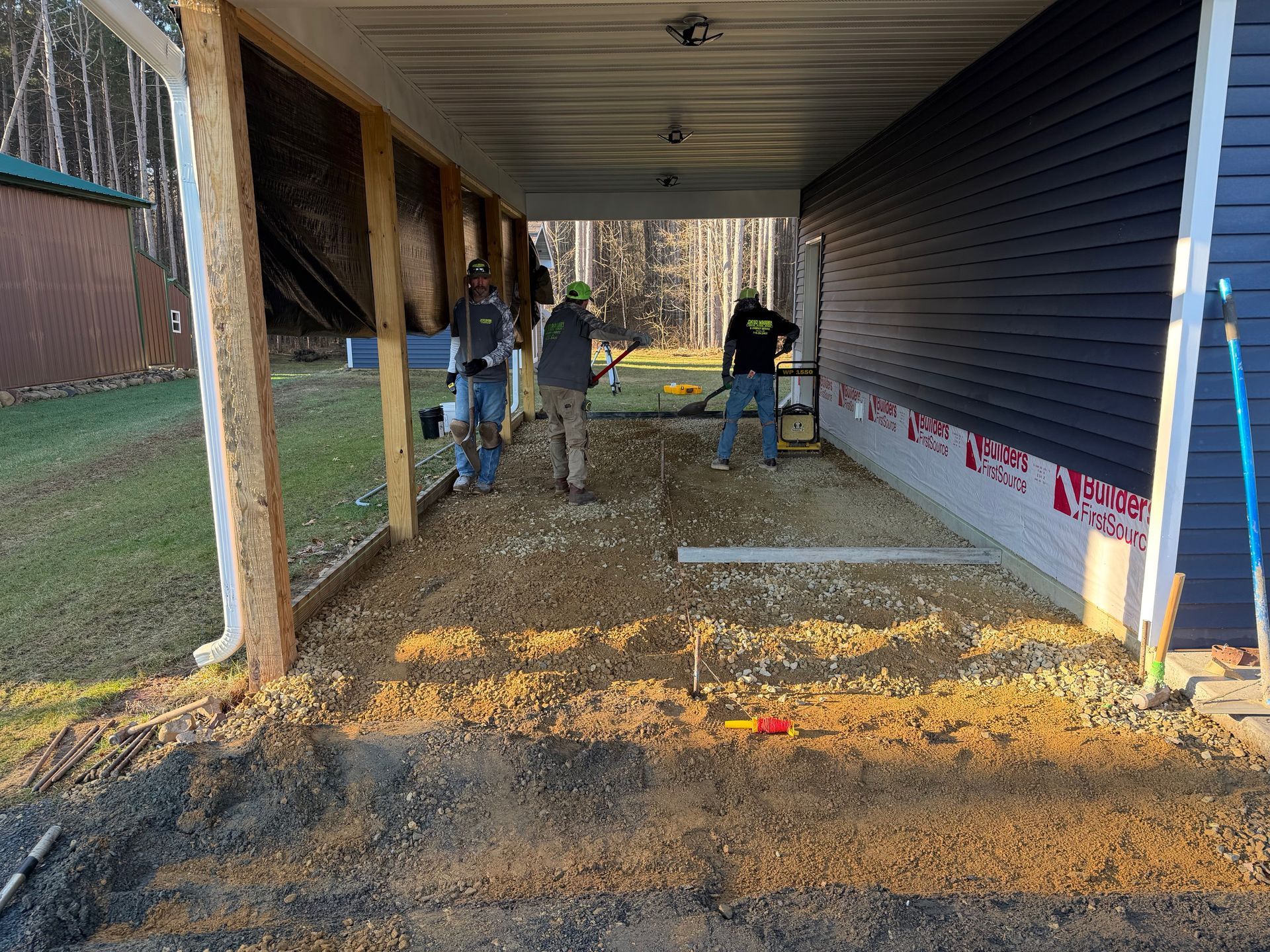 Two workers under a house frame with dirt flooring and siding being installed outdoors