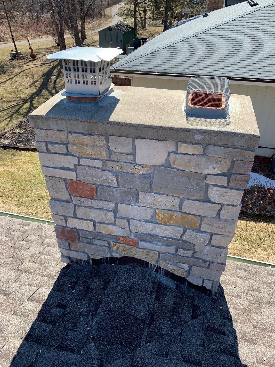 Stone chimney on a shingled roof with two metal chimney caps, seen from above outdoors.