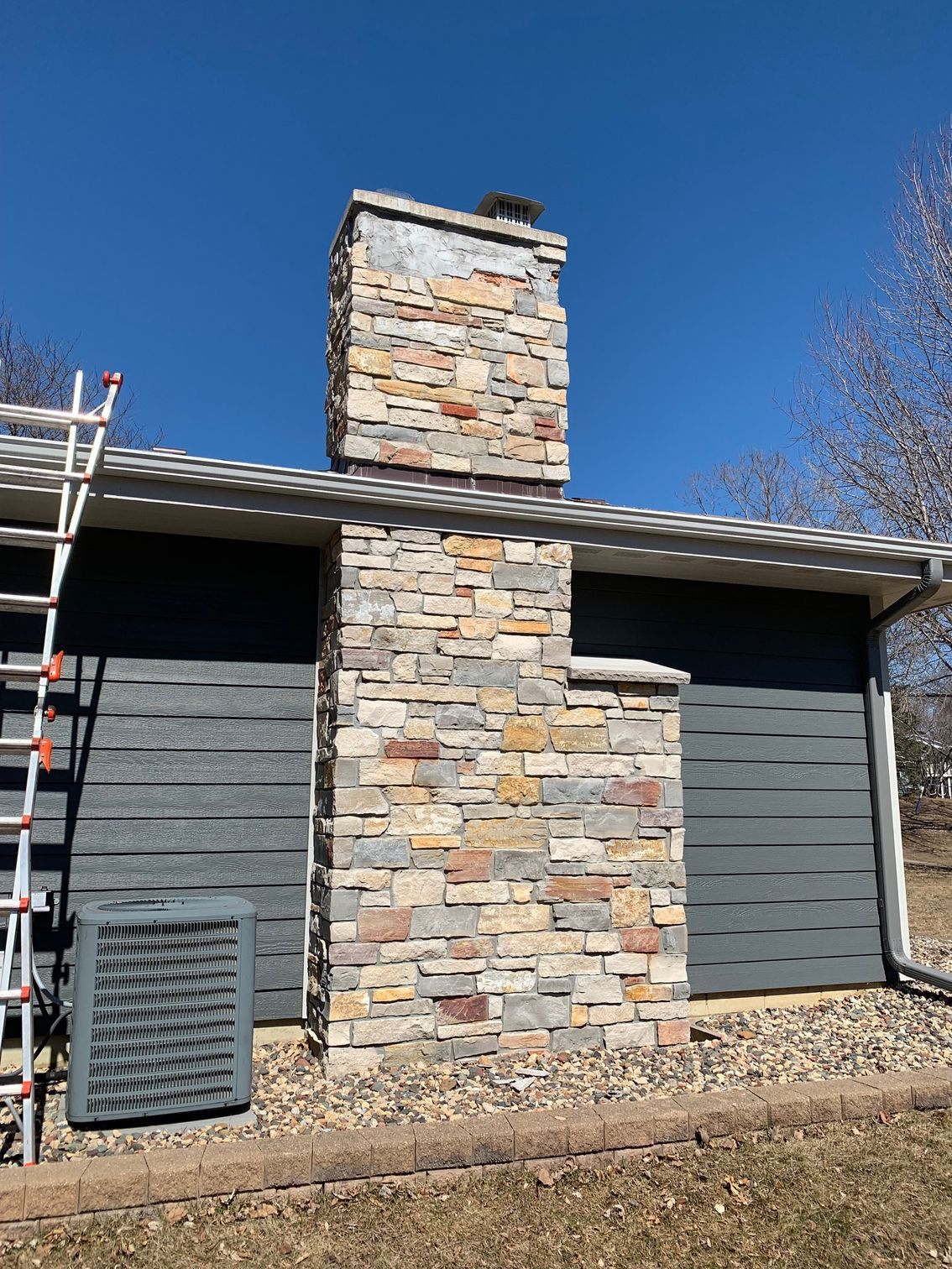 Stone chimney on a gray house exterior with a ladder at left under a clear blue sky