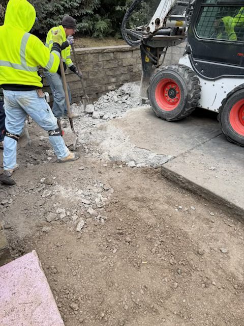Workers in high-visibility vests repair broken pavement beside a skid steer on a sidewalk.