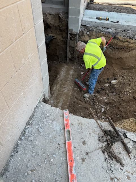 Worker in neon vest digging a trench beside a building foundation.
