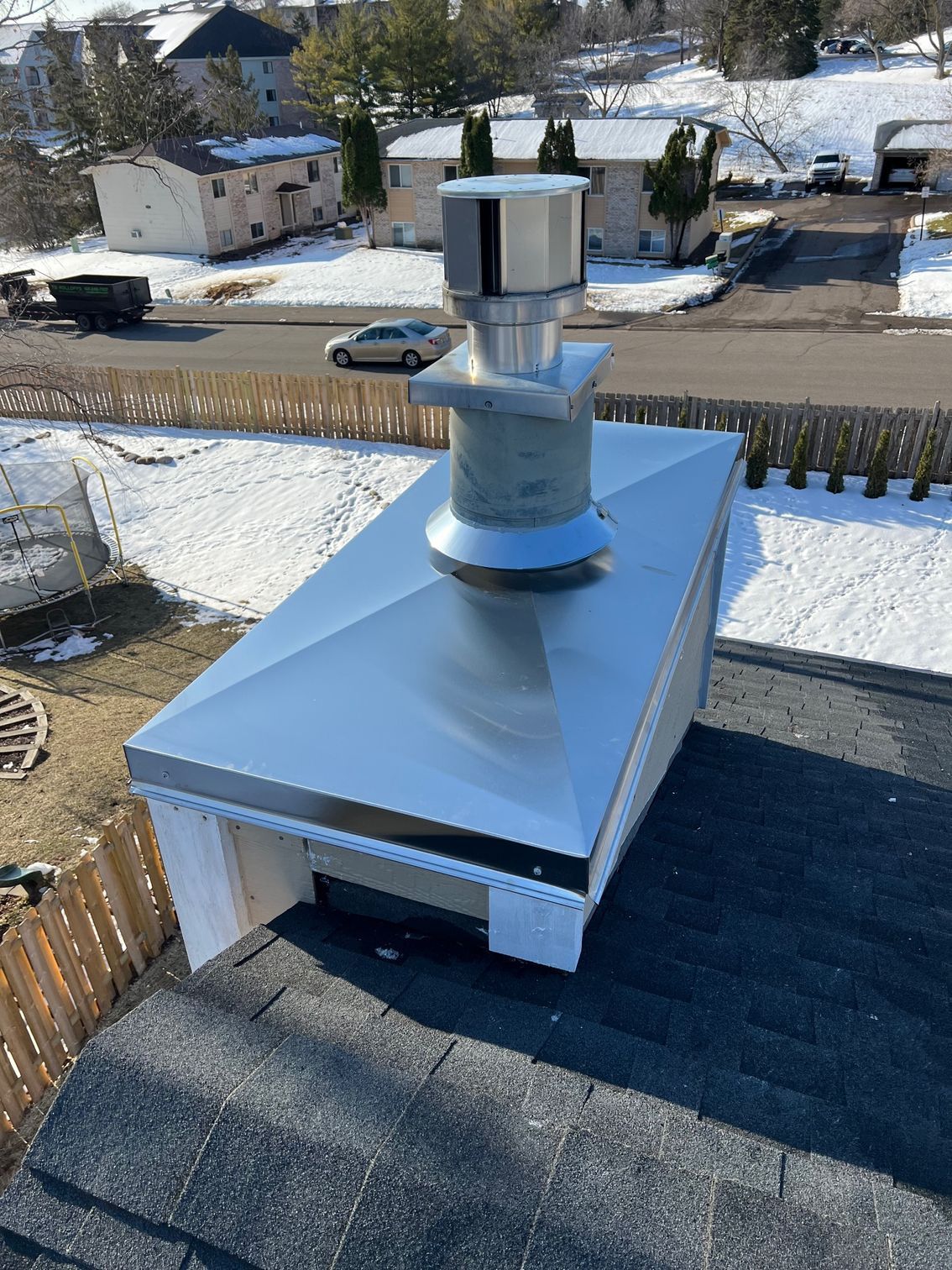 Metal roof vent on a flat rooftop beside a snowy residential street, with houses and cars in the background