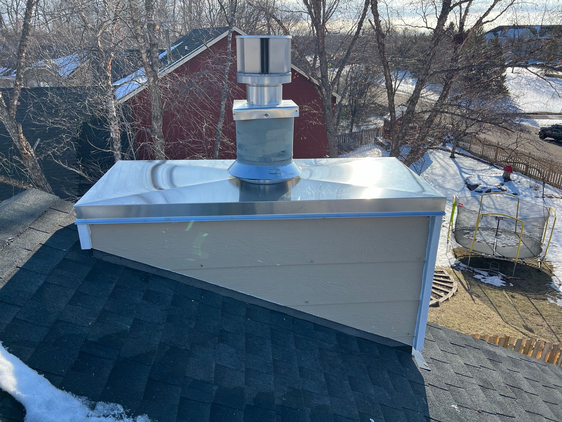 House roof with a silver chimney stack and vent, surrounded by trees and winter scenery