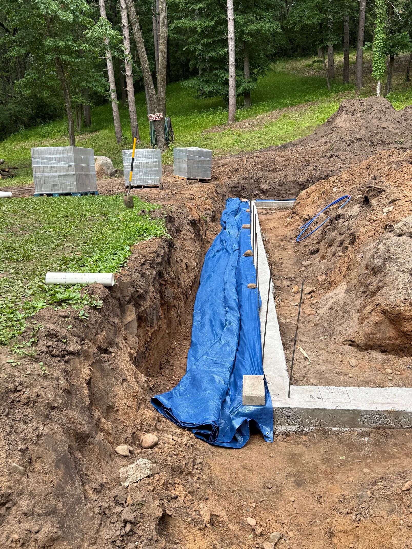 Excavation trench with blue tarp lining and concrete forms in a wooded outdoor area