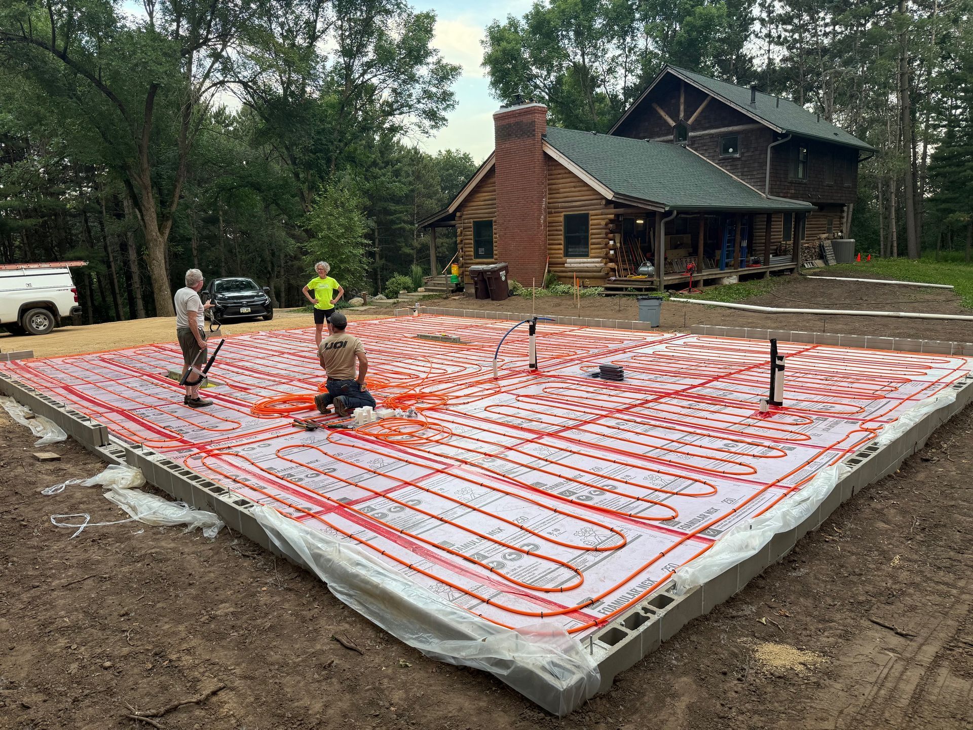 Workers installing red radiant floor tubing on a concrete slab beside a house.