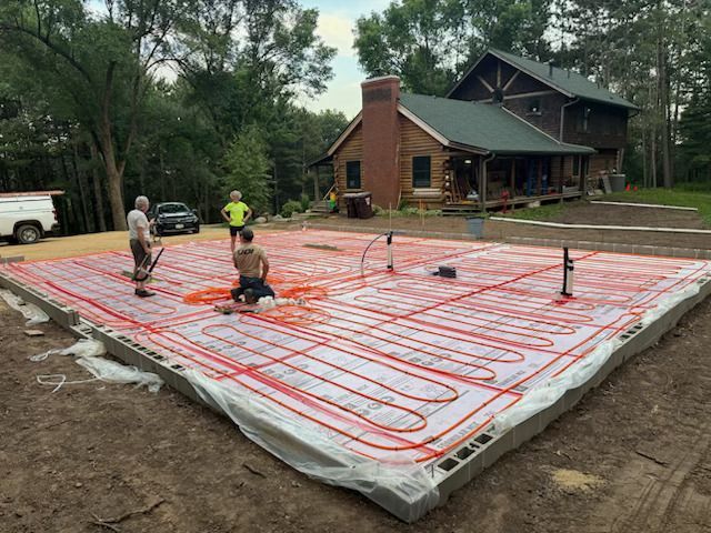 Workers installing red radiant floor tubing on a concrete slab outside a house under construction.