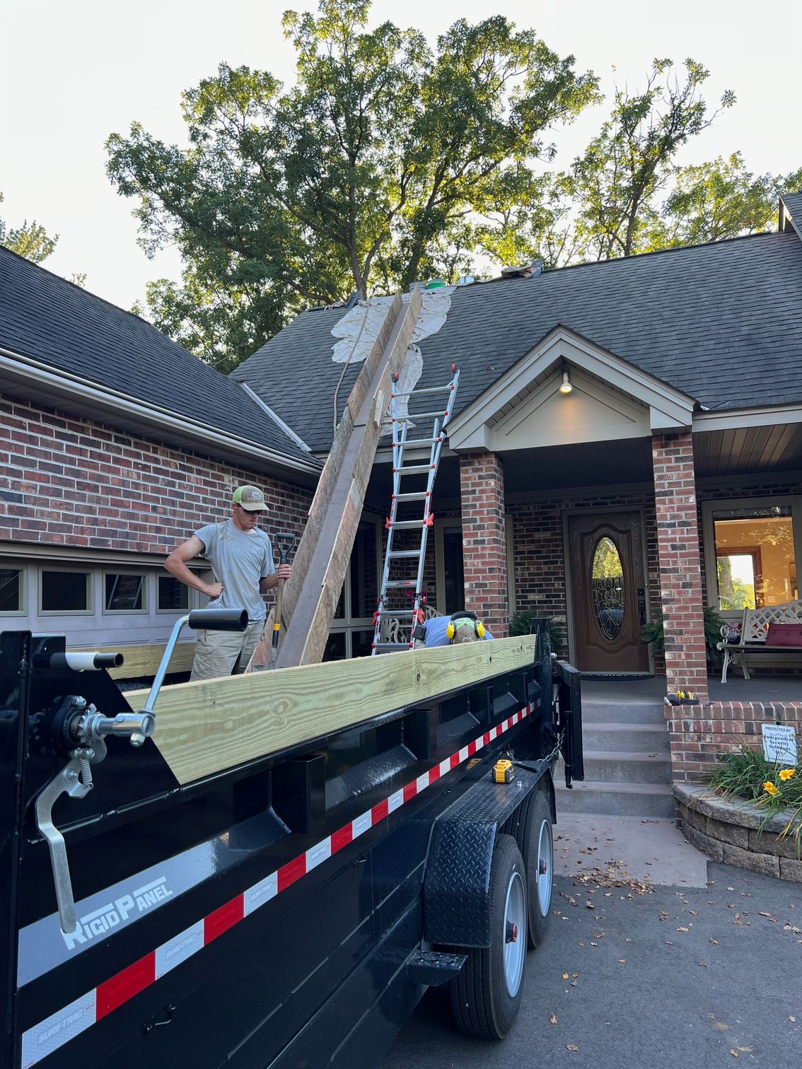 Workers unloading a ladder from a trailer beside a brick house with a ladder propped to the roof