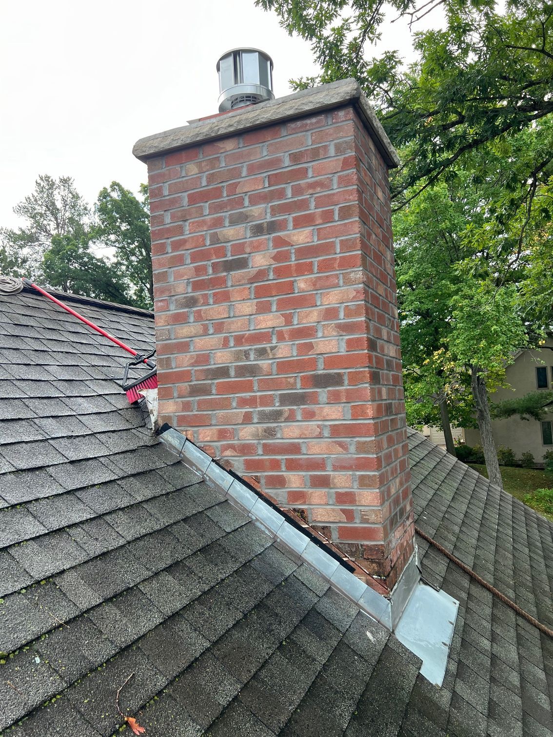 Brick chimney on a gray shingled roof with a metal cap, surrounded by trees.
