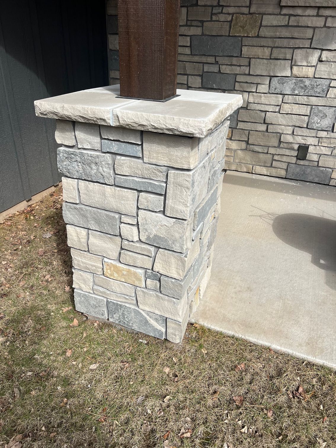 Stone porch column beside a concrete walkway and grass, supporting a wooden post.