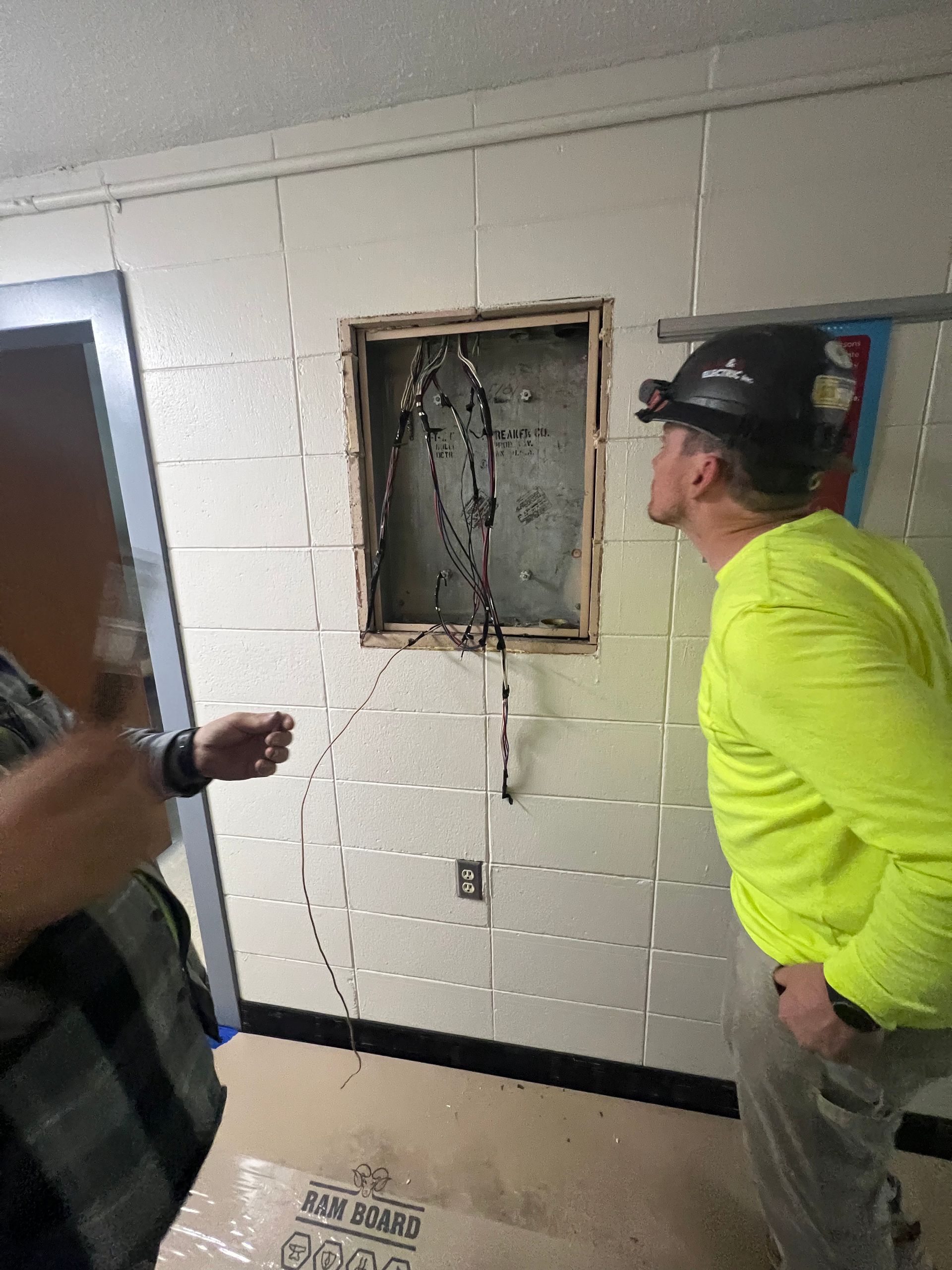 Two people inspect an open wall panel in a white cinderblock hallway.