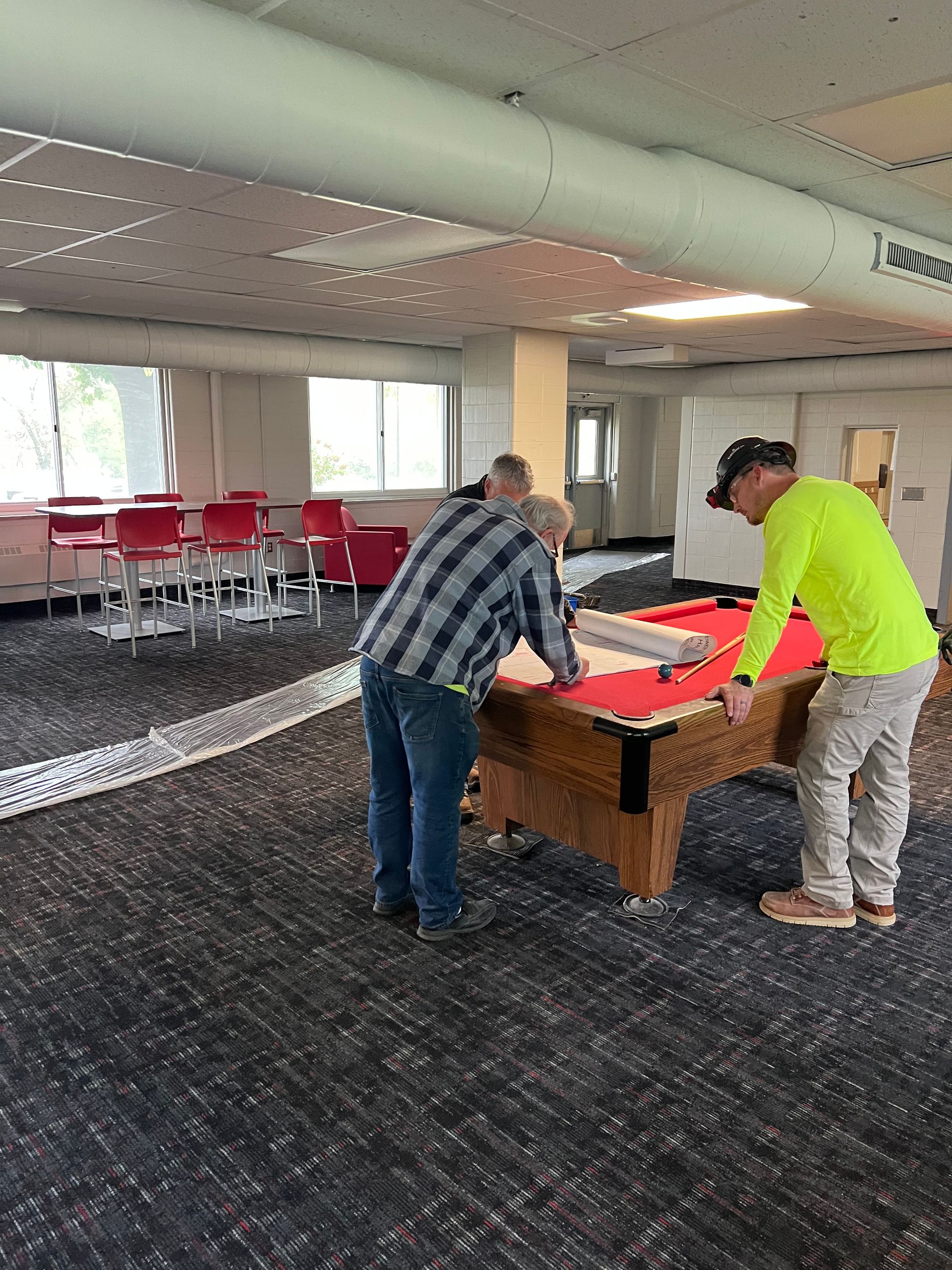 Two people setting up a red table tennis table in a carpeted lounge with rows of red chairs.