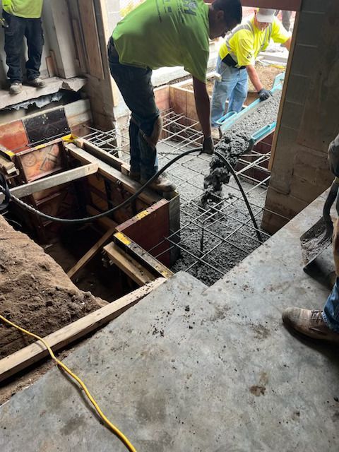 Workers pouring concrete into a floor opening at a construction site, with wooden formwork and tools visible.