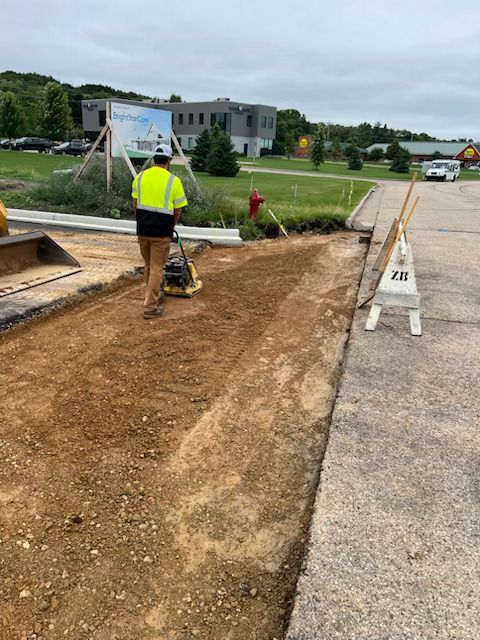 Worker compacts dirt beside a curb on a roadway construction site under overcast skies.
