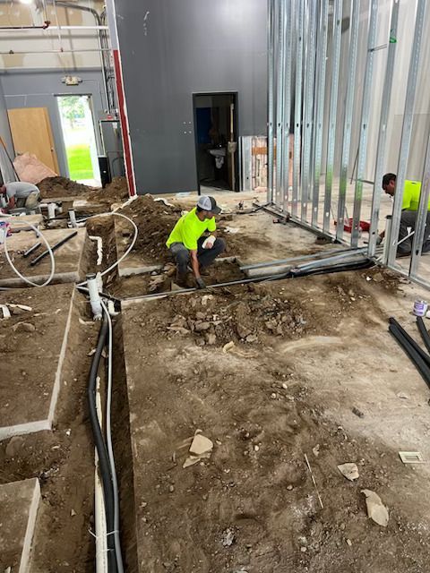Construction worker in neon yellow shirt kneeling in a muddy interior renovation site with exposed framing and hoses