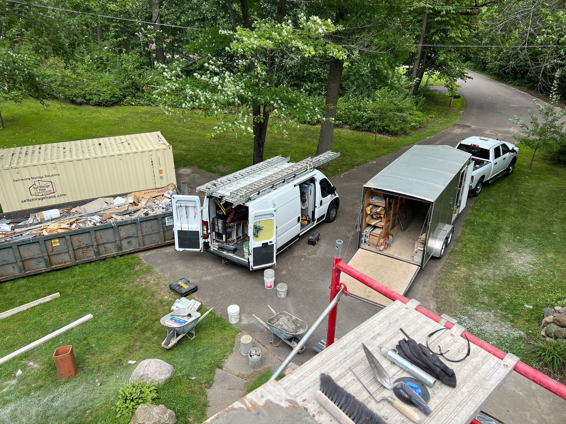 Two work vans and a trailer beside a yellow shed in a wooded yard, with tools and materials on the ground.
