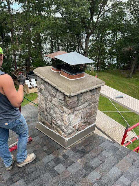Stone chimney on a shingled roof, with a person standing nearby and trees in the background.